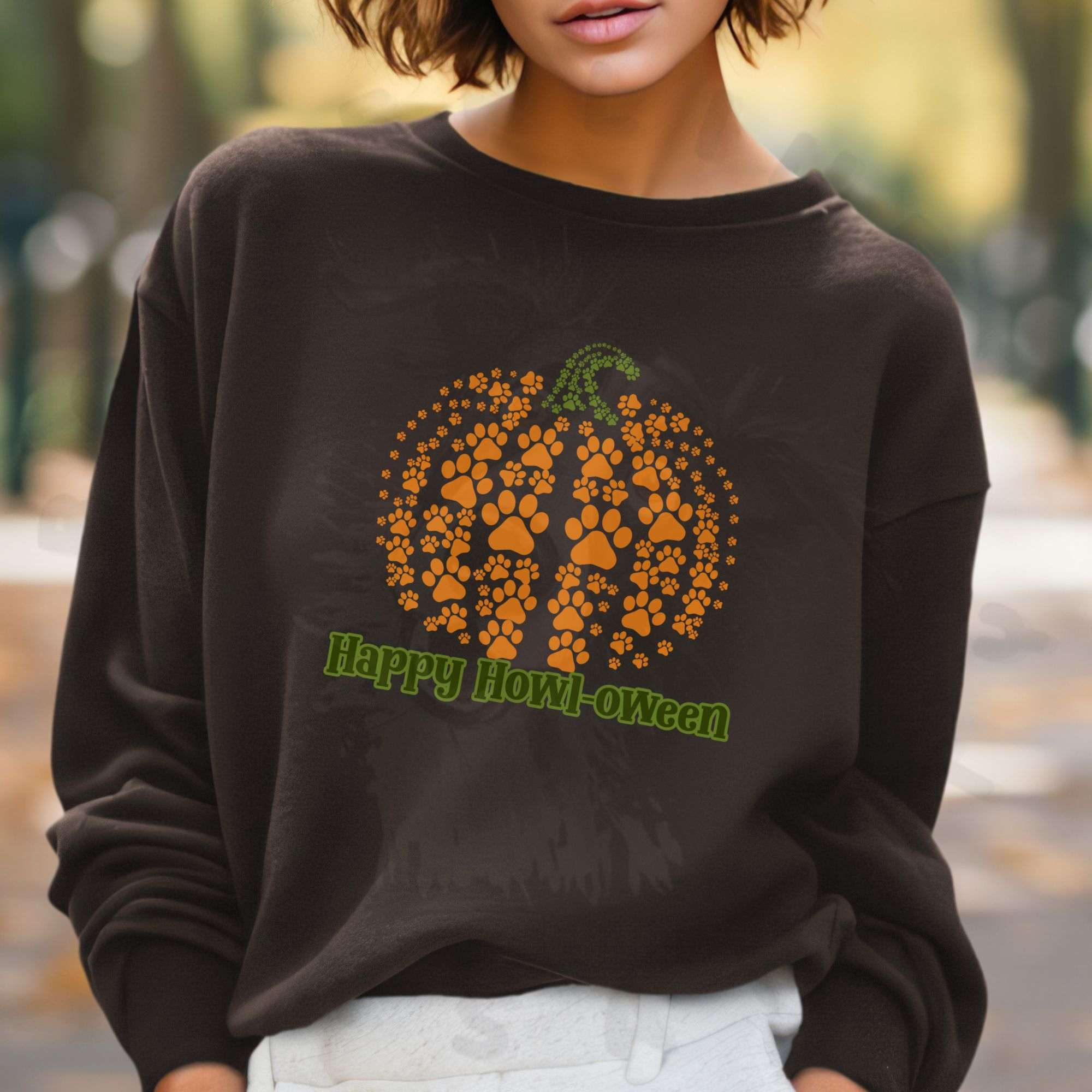 a woman standing in a park wearing a brown halloween sweatshirt that says happy howl-oween beneath a pumpkin made of pawprints