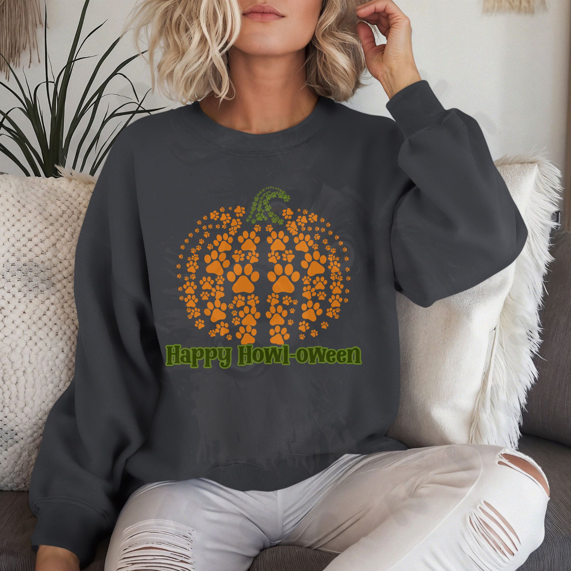 a woman sitting on a white sofa wearing a charcoal gray halloween sweatshirt that says happy howl-oween with a pumpkin made of pawprints above it