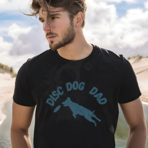 a man standing on a beach wearing a black disc dog dad t-shirt featuring a silhouette of a dog leaping for a frisbee