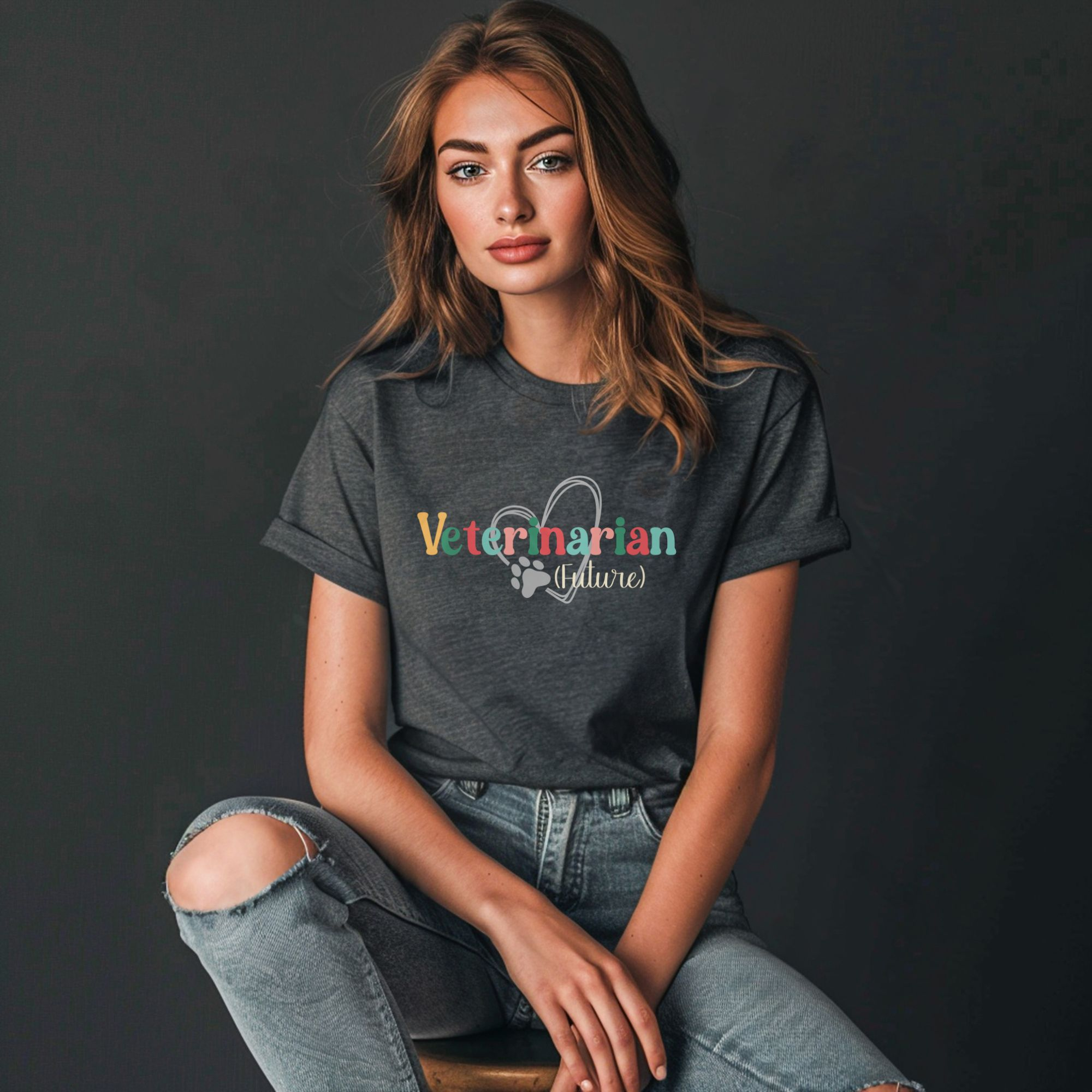 a woman sitting on a stool in front of a dark gray background wearing ripped jeans and a dark gray heather future veterinarian tee with a heart and paw print