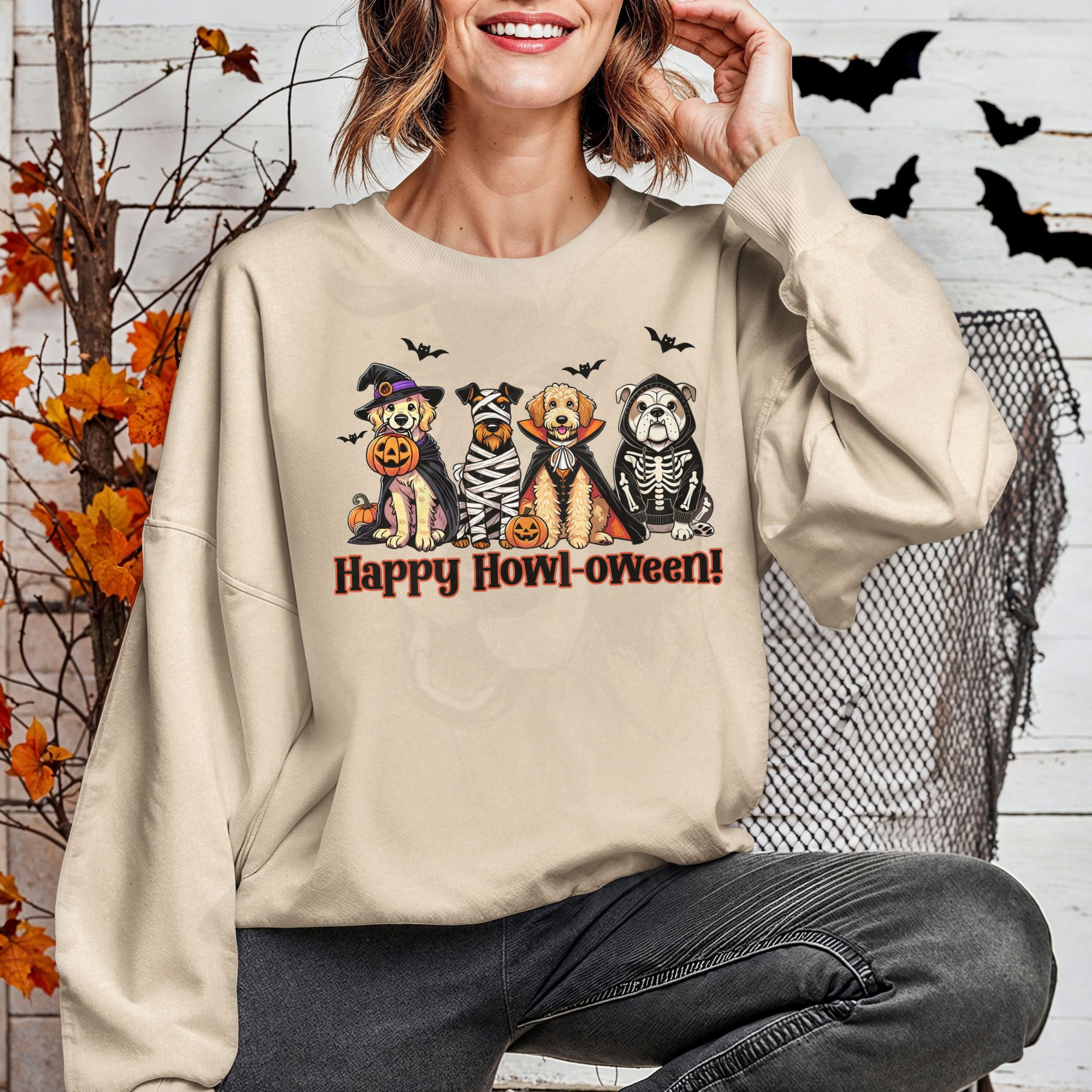 a woman in front of a halloween background wearing a sand sweatshirt featuring a graphic of 4 dogs wearing halloween costumes and the phrase happy howl-oween
