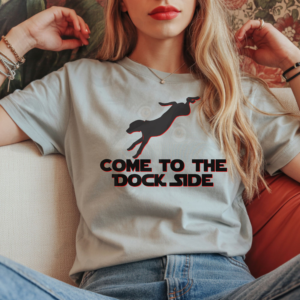 a long-haired woman leaning back on a sofa wearing a silver come to the dock side tee with a silhouette of a diving dog