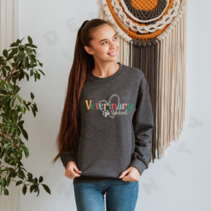 a woman standing in front of a boho background wearing a dark gray heather sweatshirt with a colorful veterinary student design with a heart and pawprint