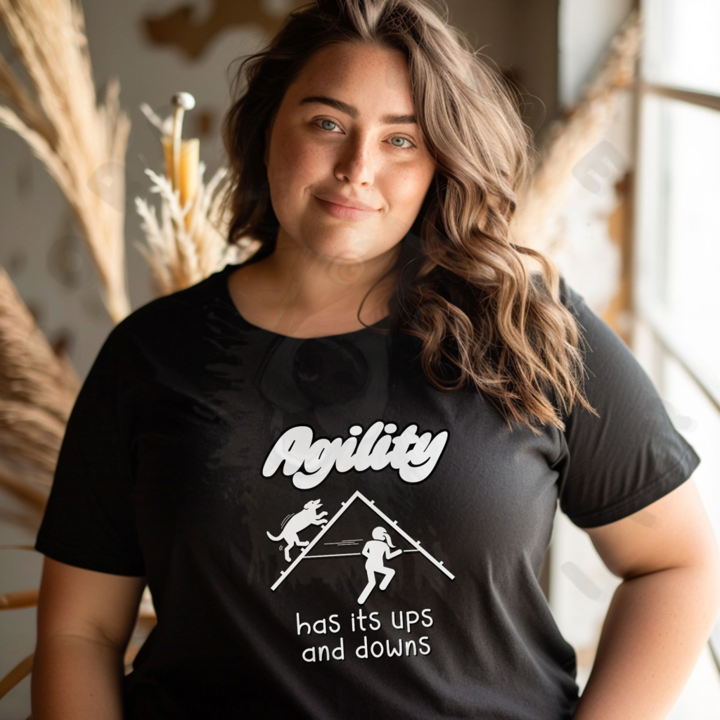 a woman standing in front of a boho background wearing a heather team purple agility has its ups and downs tee with a graphic of a dog climbing an agility ramp