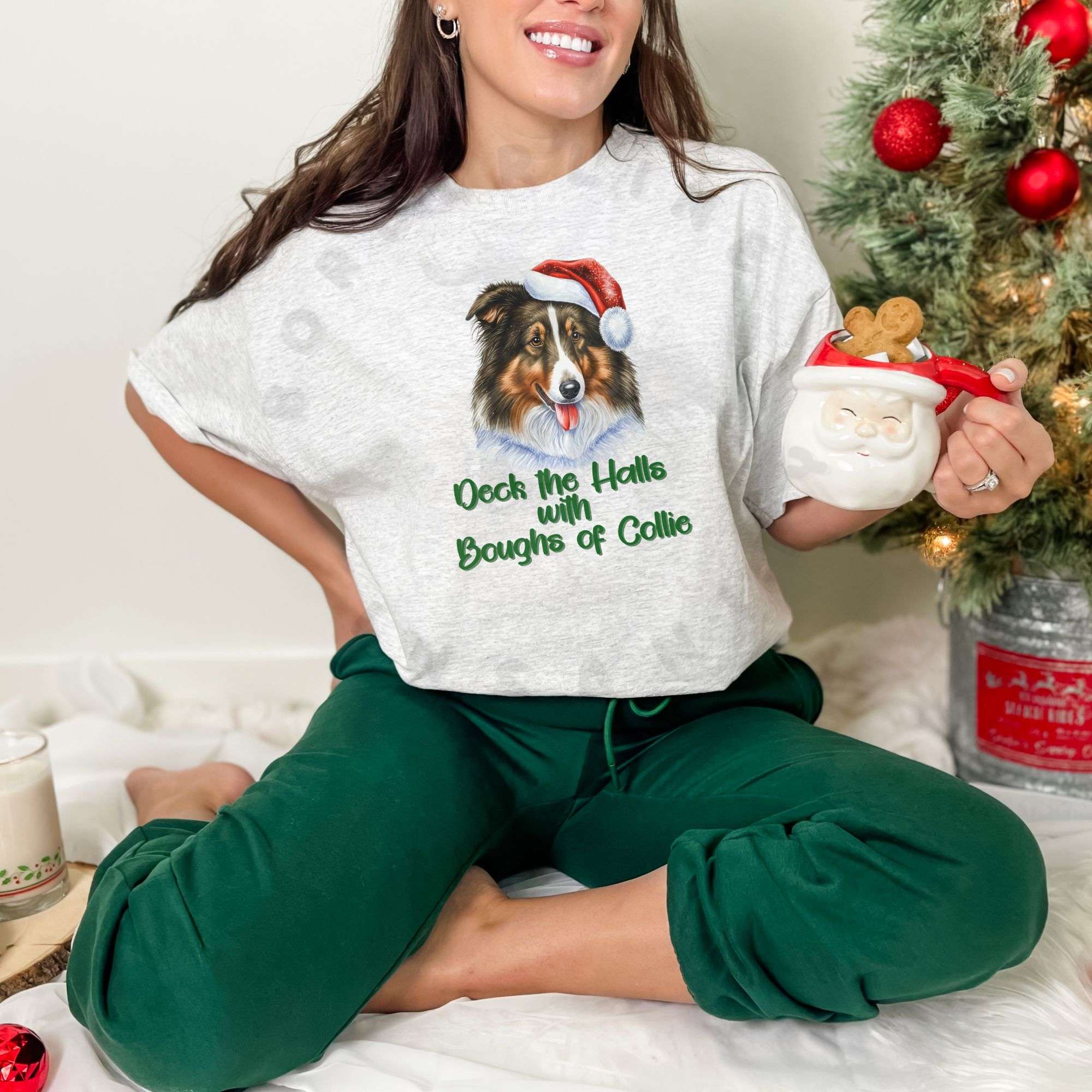 a woman holding a santa mug and sitting next to a christmas tree wearing an ash grey tee that says deck the halls with boughs of collie under a rough collie wearing a santa hat
