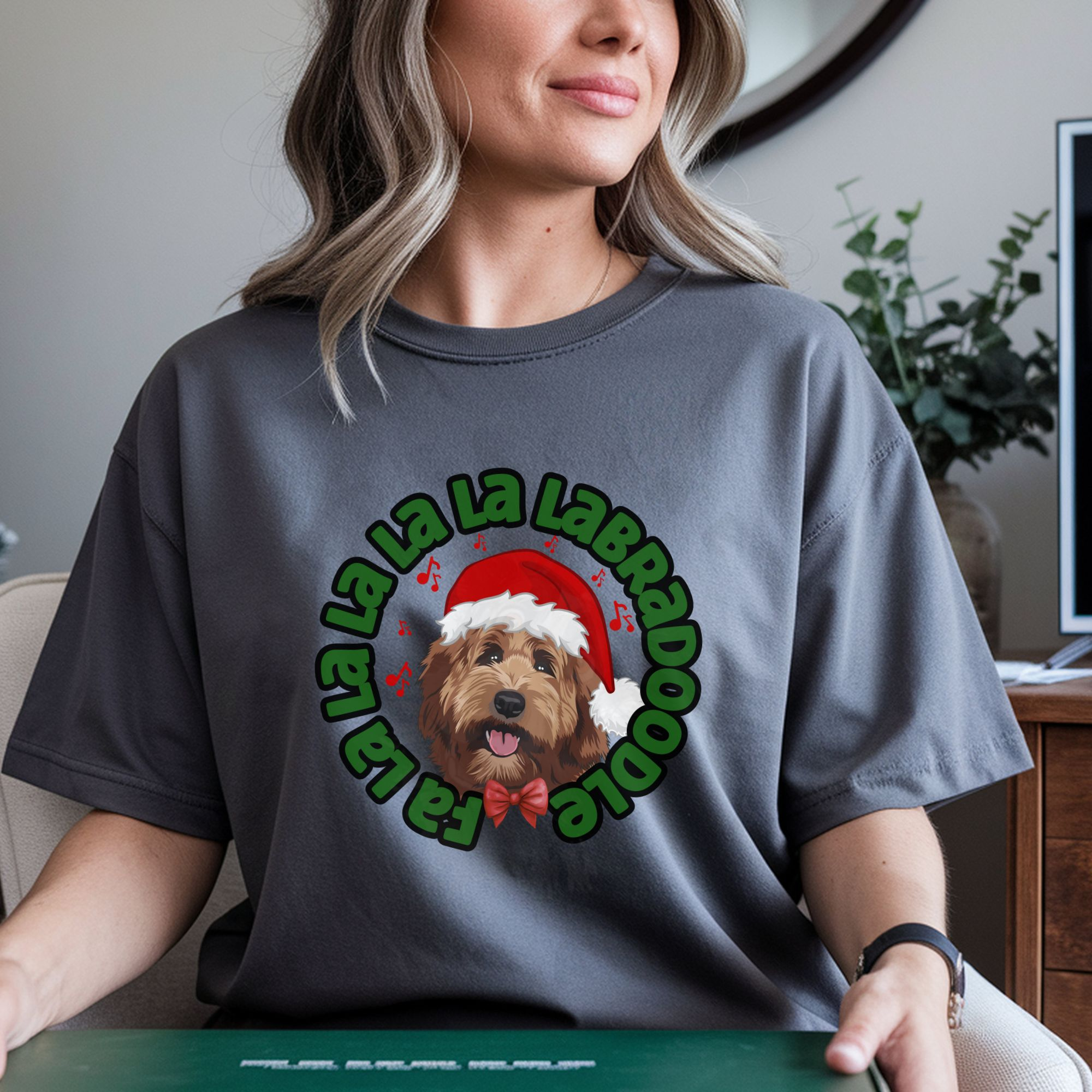 a woman sitting in front of a plant wearing an asphalt grey tee featuring a graphic of a labradoodle wearing a santa hat and the words Fa la la la la la labradoodle