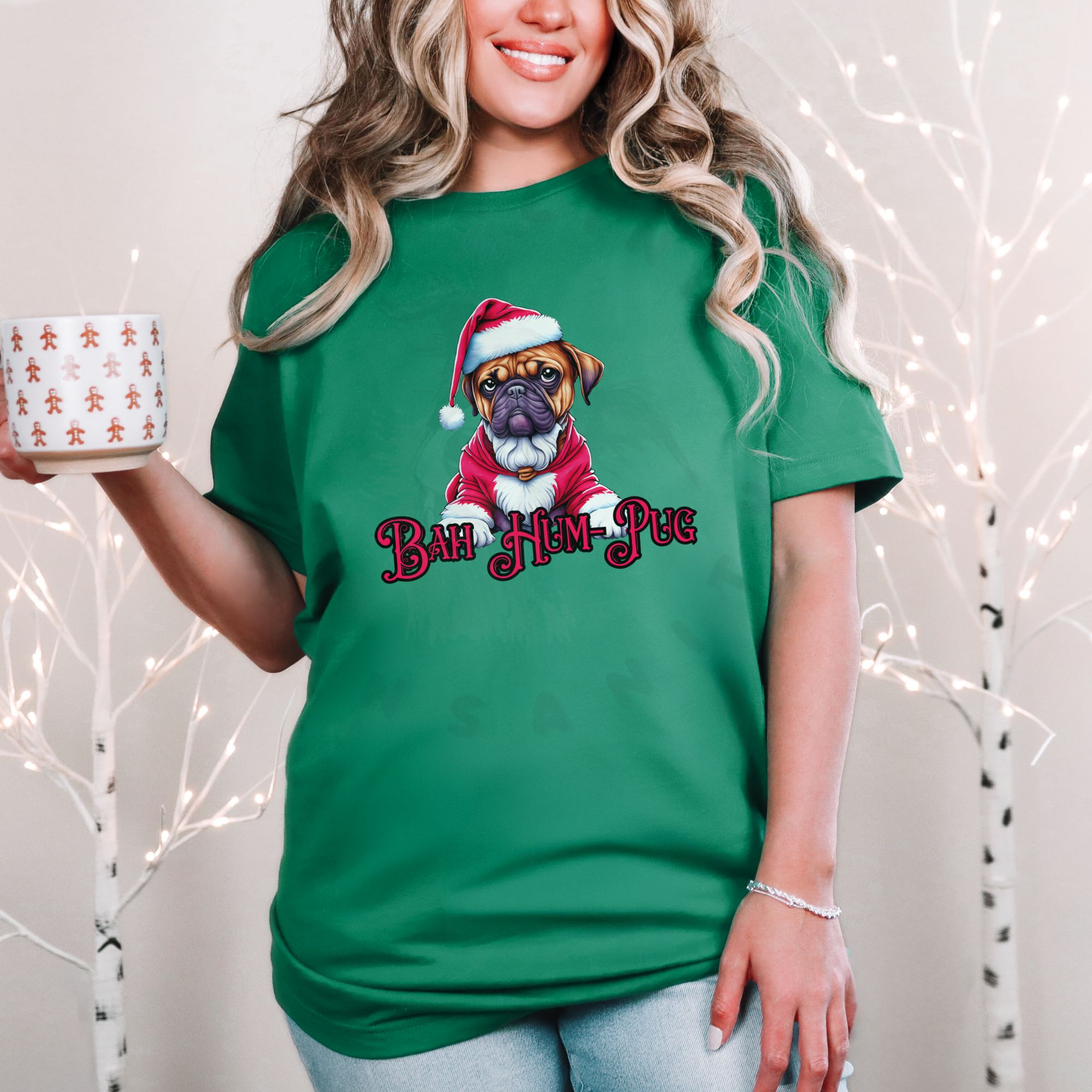 a woman standing in front of a christmas background holding a holiday mug wearing a heather kelly green t-shirt that says bah hum-pug below a graphic of a grumpy pug wearing a santa suit