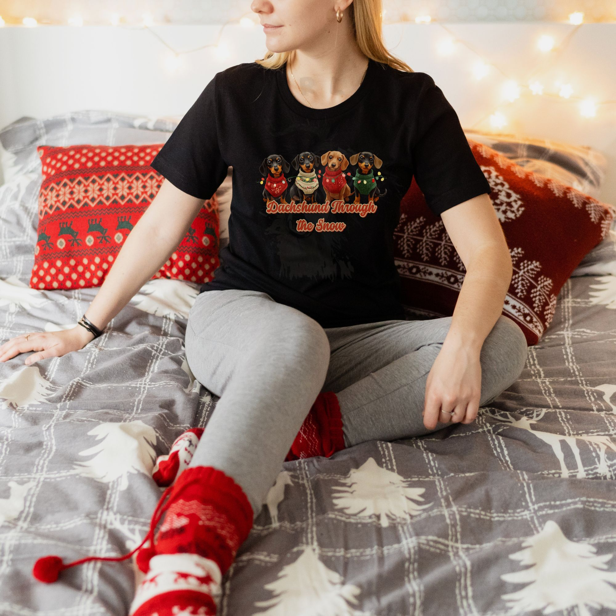 a woman sitting against christmas pillows wearing a black dachshund through the snow t-shirt featuring 4 dachshunds wearing christmas sweaters and tangled in christmas lights