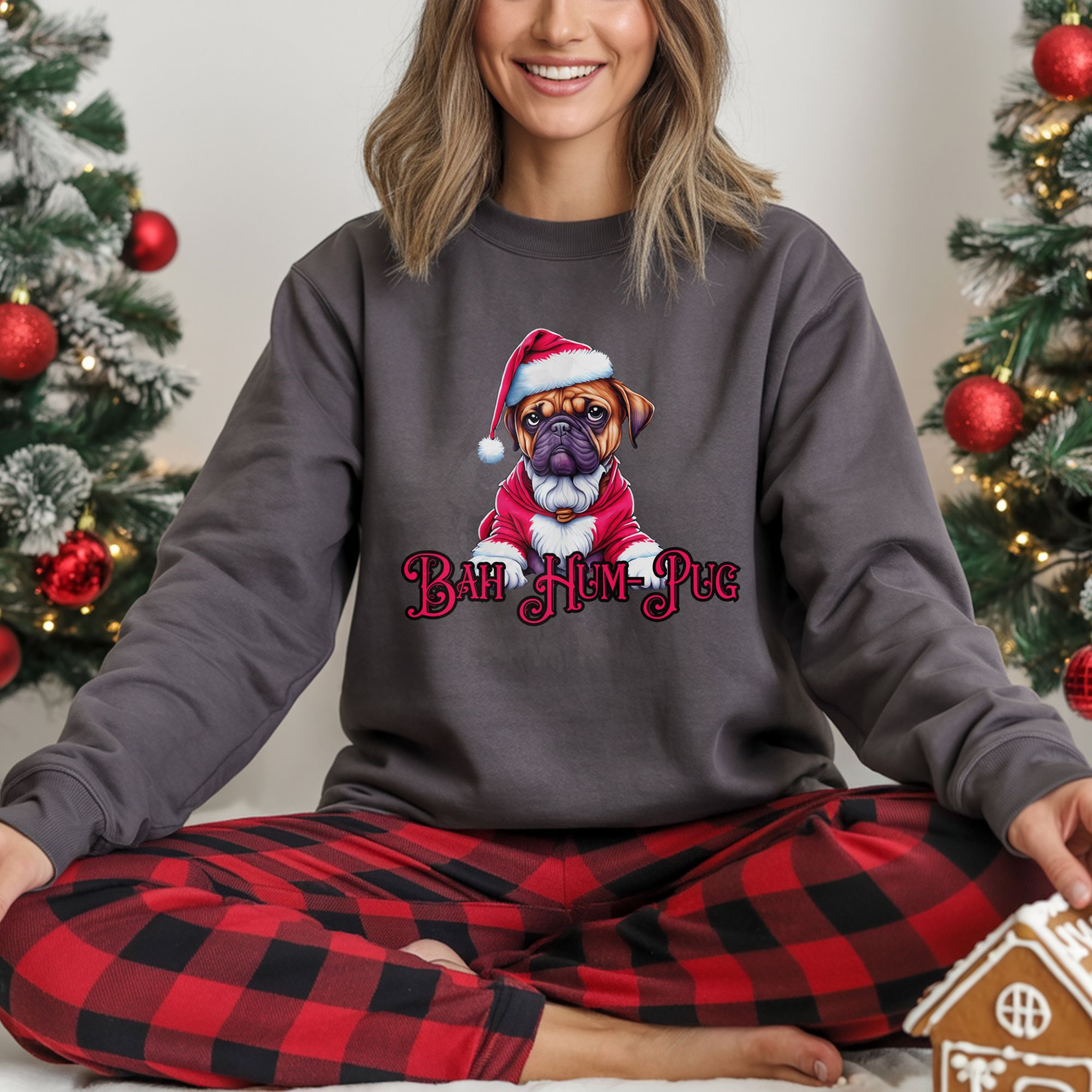 a woman sitting between two christmas trees wearing a charcoal sweatshirt that says bah hum-pug below a graphic of a grumpy pug wearing a santa suit