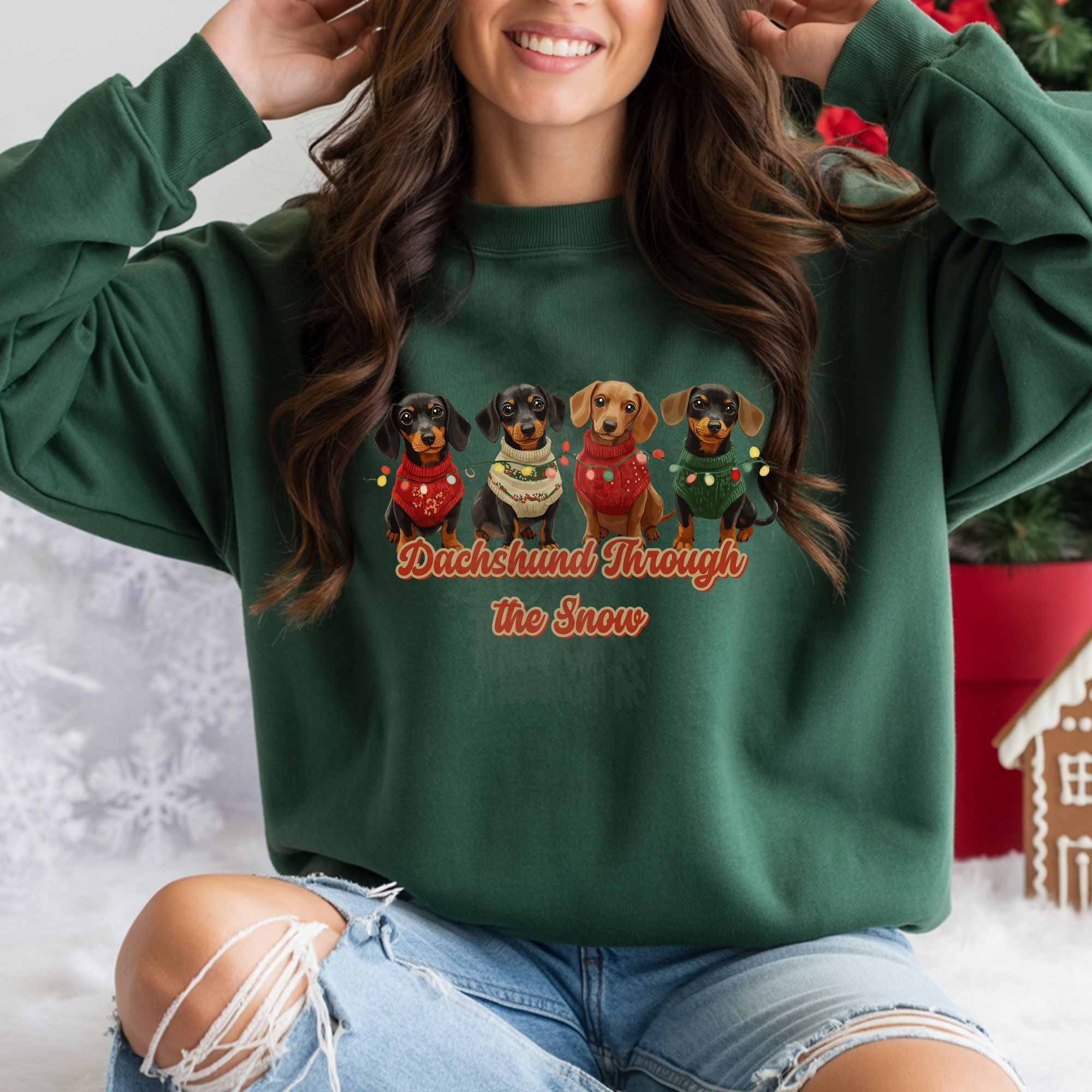 a woman sitting in front of a gingerbread house and other decorations wearing a forest dachshund through the snow sweatshirt featuring 4 dachshunds wearing christmas sweaters and tangled in christmas lights