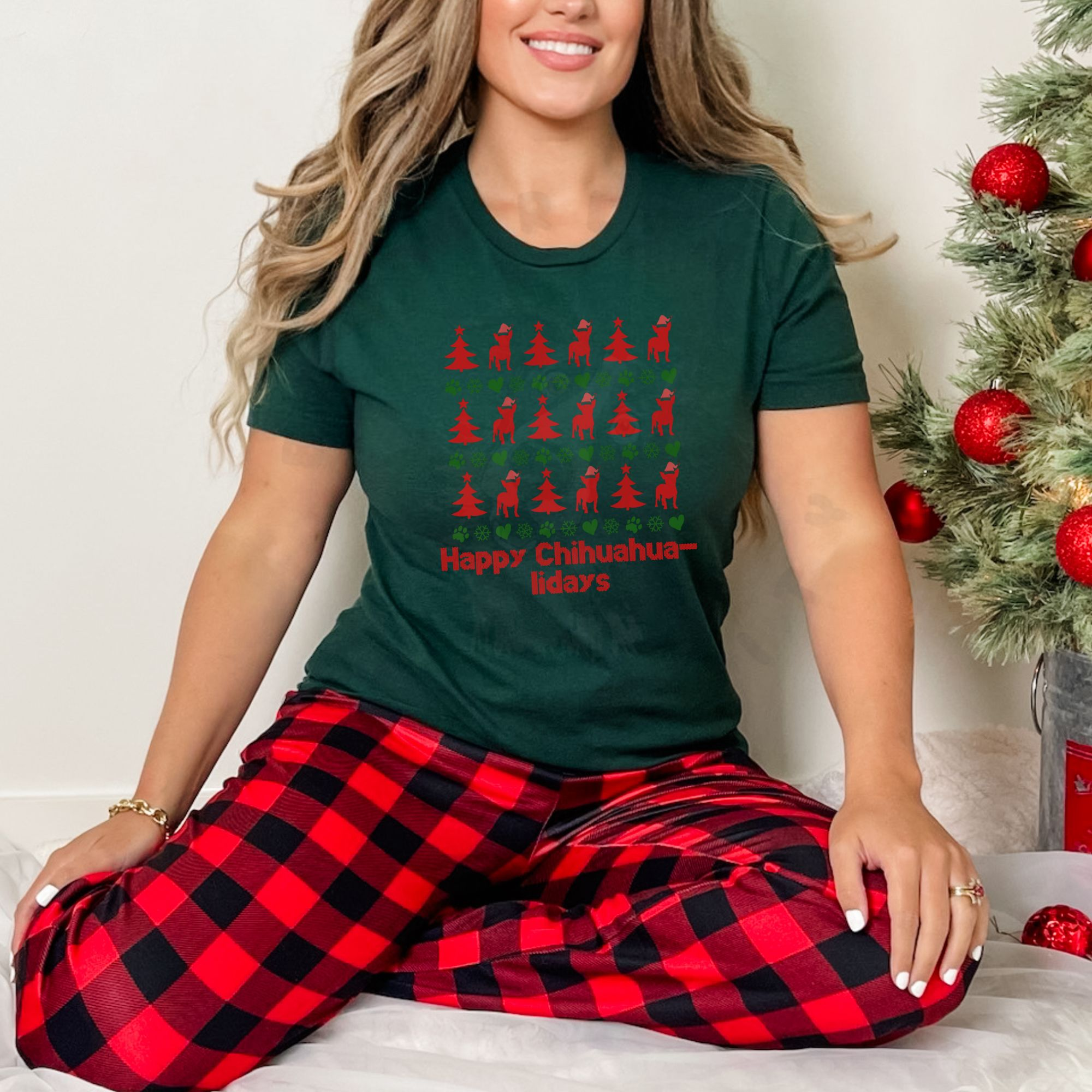 a woman in red buffalo plaid pajama pants sitting next to a christmas tree wearing a forest green t-shirt designed ugly christmas sweater style with rows of red christmas trees, green snowflakes, and chihuahuas wearing santa hats.