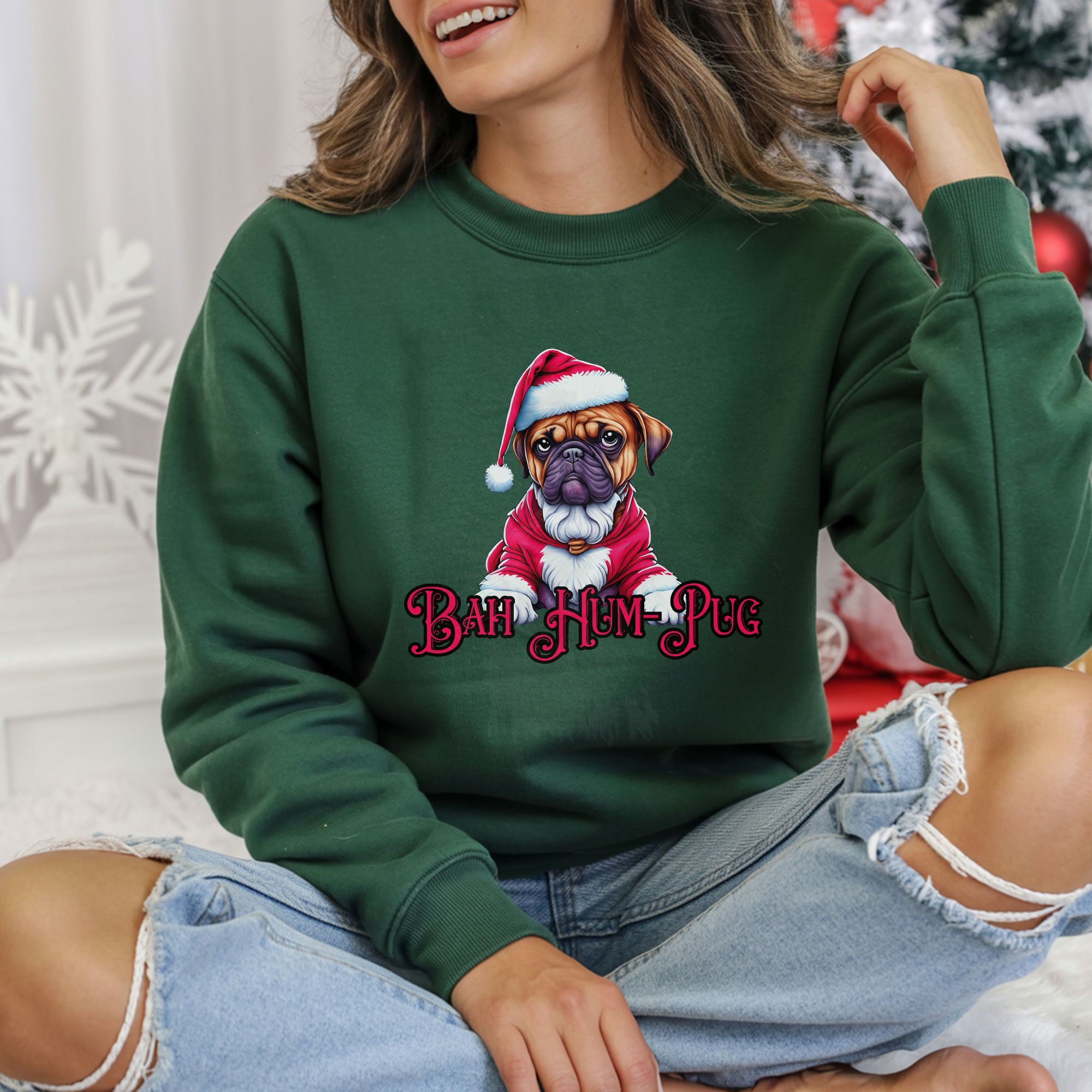 a woman sitting in front of a christmas tree wearing a forest green sweatshirt that says bah hum-pug below a graphic of a grumpy pug wearing a santa suit
