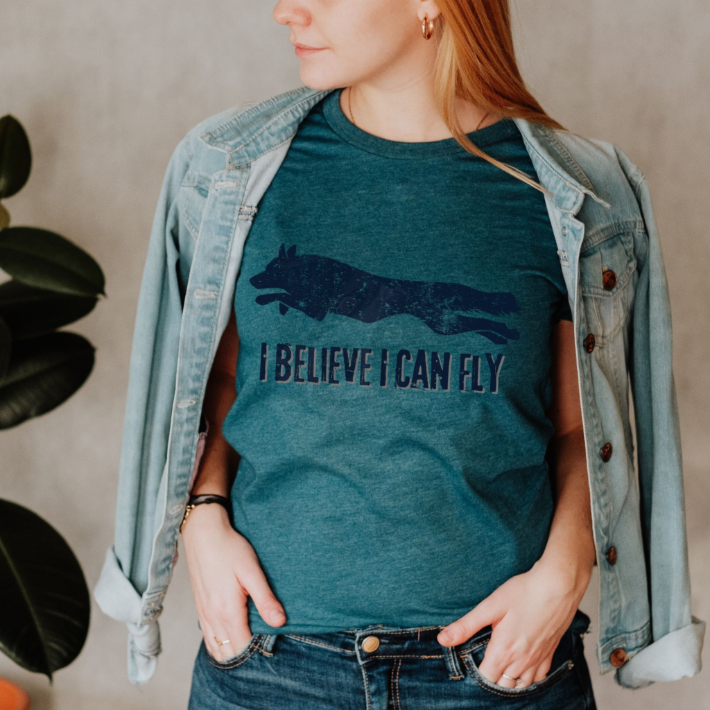 a woman with a denim jacket wrapped around her shoulders wearing a heather deep teal tee with a silhouette of a leaping dog that ways i believe i can fly below it