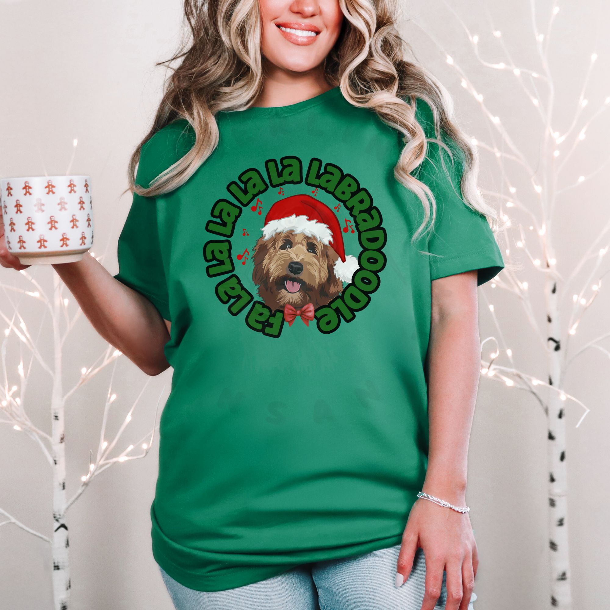 a woman standing in front of a christmas background holding a holiday mug wearing a kelly green tee featuring a graphic of a labradoodle wearing a santa hat and the words Fa la la la la la labradoodle