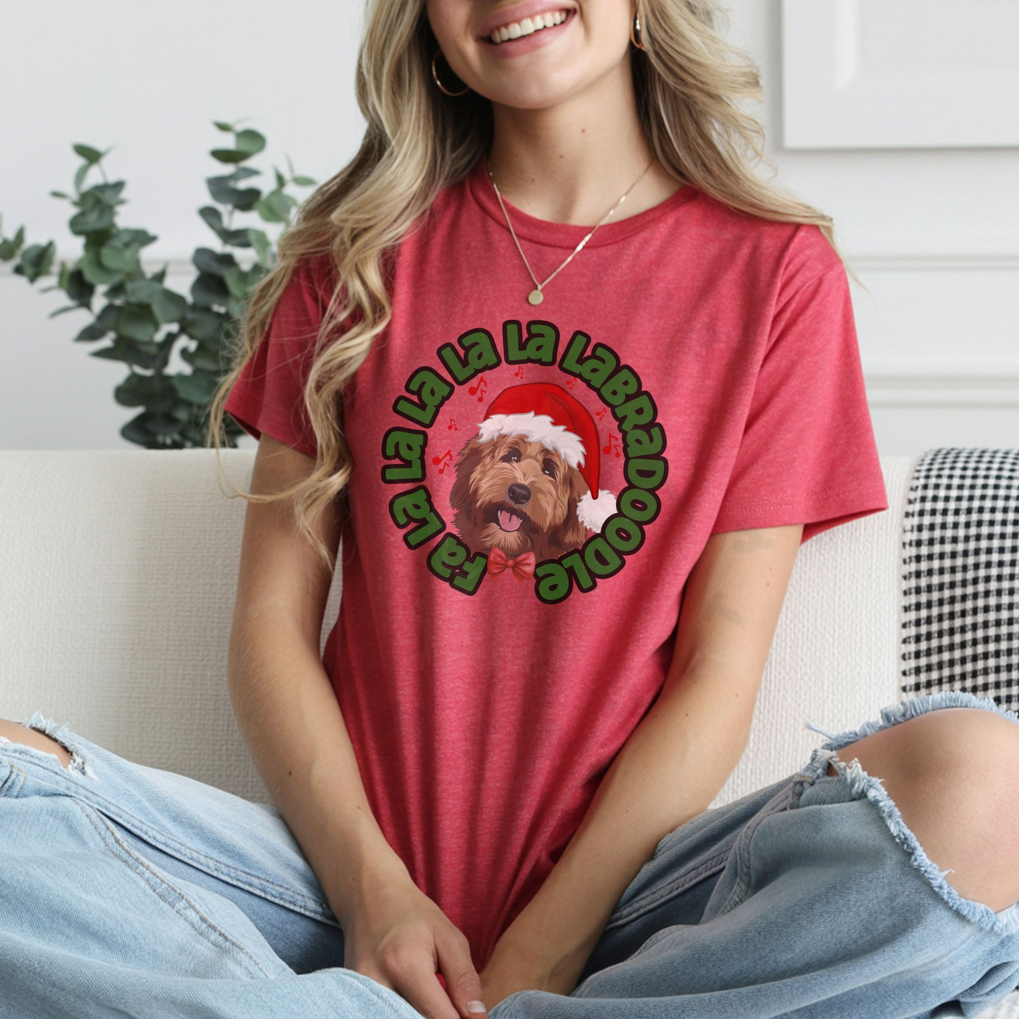 a woman wearing ripped jeans sitting on a couch wearing a heather red tee featuring a graphic of a labradoodle wearing a santa hat and the words Fa la la la la la labradoodle