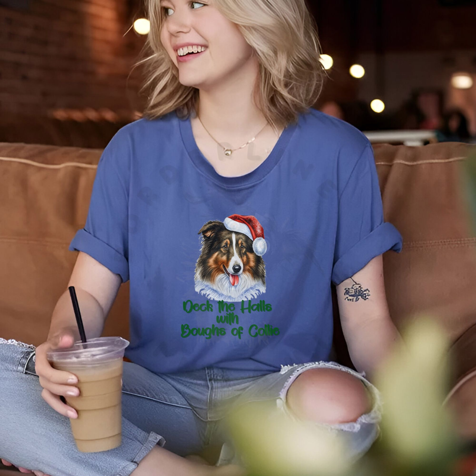 a woman sitting in a coffee shop boothe holding an iced coffee wearing a heather true royal tee that says deck the halls with boughs of collie in cursive with a rough collie wearing a santa hat