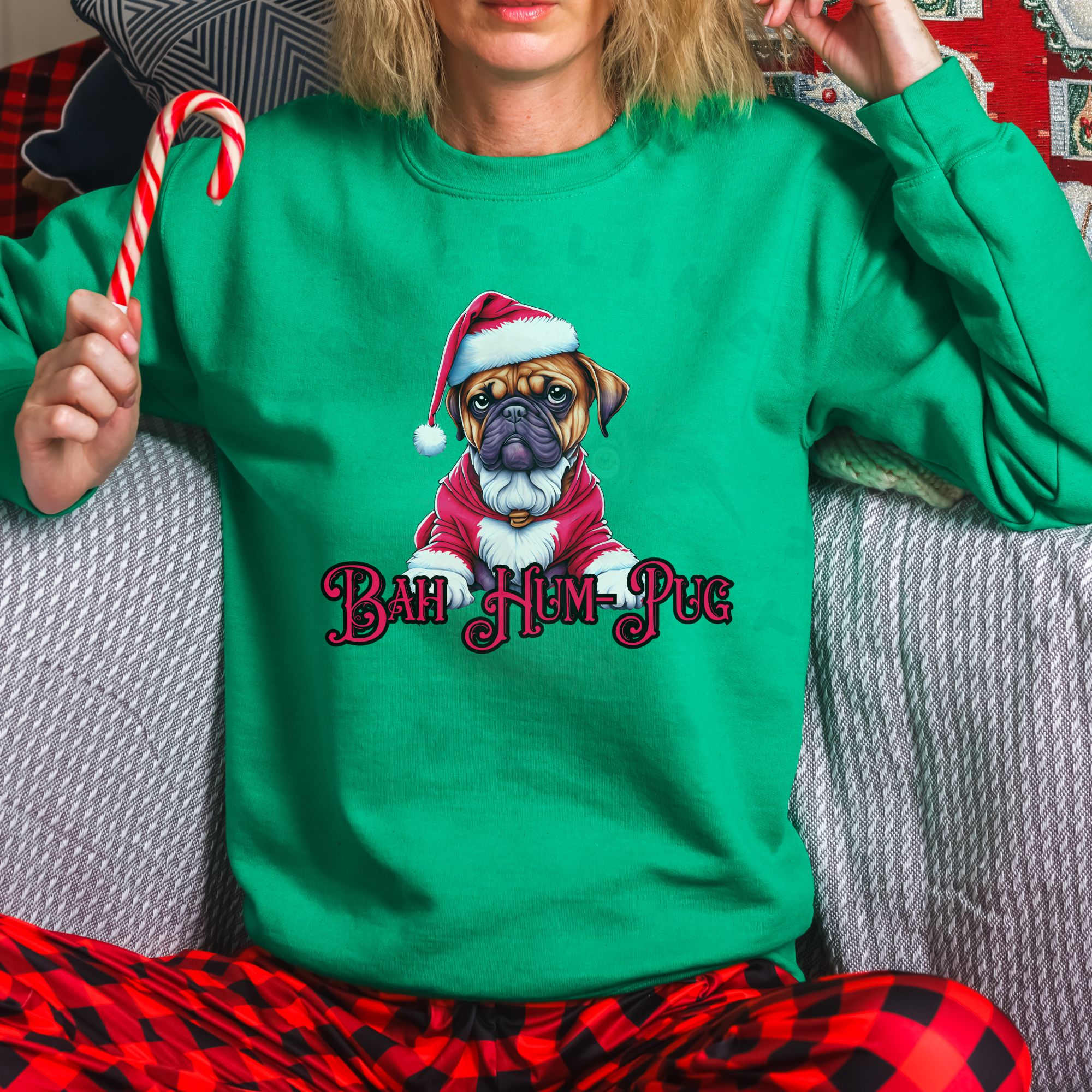 a woman sitting on a sofa holding a candy cane wearing an irish green sweatshirt that says bah hum-pug below a graphic of a grumpy pug wearing a santa suit