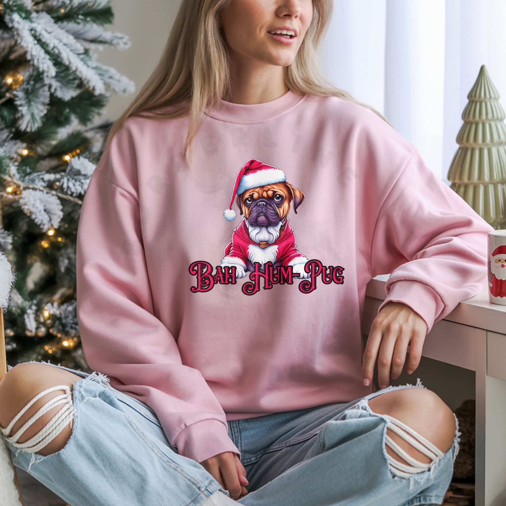 a woman sitting next to a christmas tree wearing a light pink sweatshirt that says bah hum-pug below a graphic of a grumpy pug wearing a santa suit