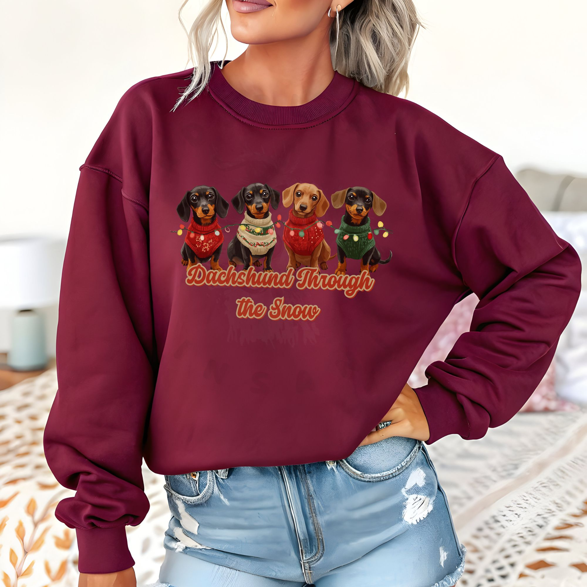a woman standing in front of a bed wearing a maroon dachshund through the snow sweatshirt featuring 4 dachshunds wearing christmas sweaters and tangled in christmas lights