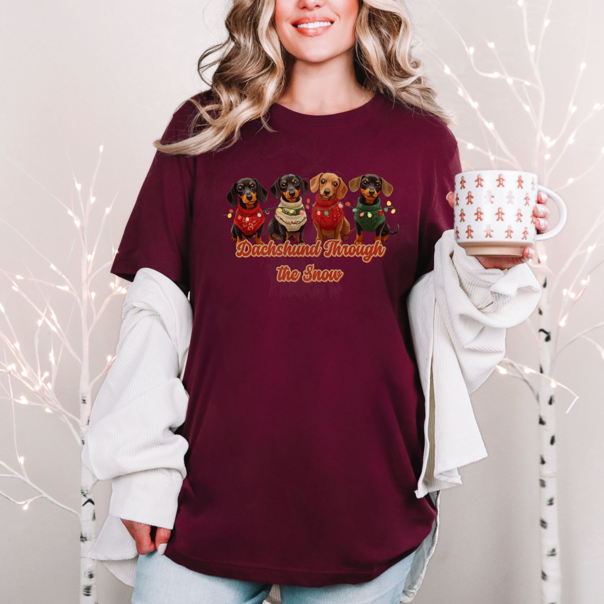 a woman standing in front of a christmas background holding a festive mug wearing a maroon dachshund through the snow t-shirt featuring 4 dachshunds wearing christmas sweaters and tangled in christmas lights