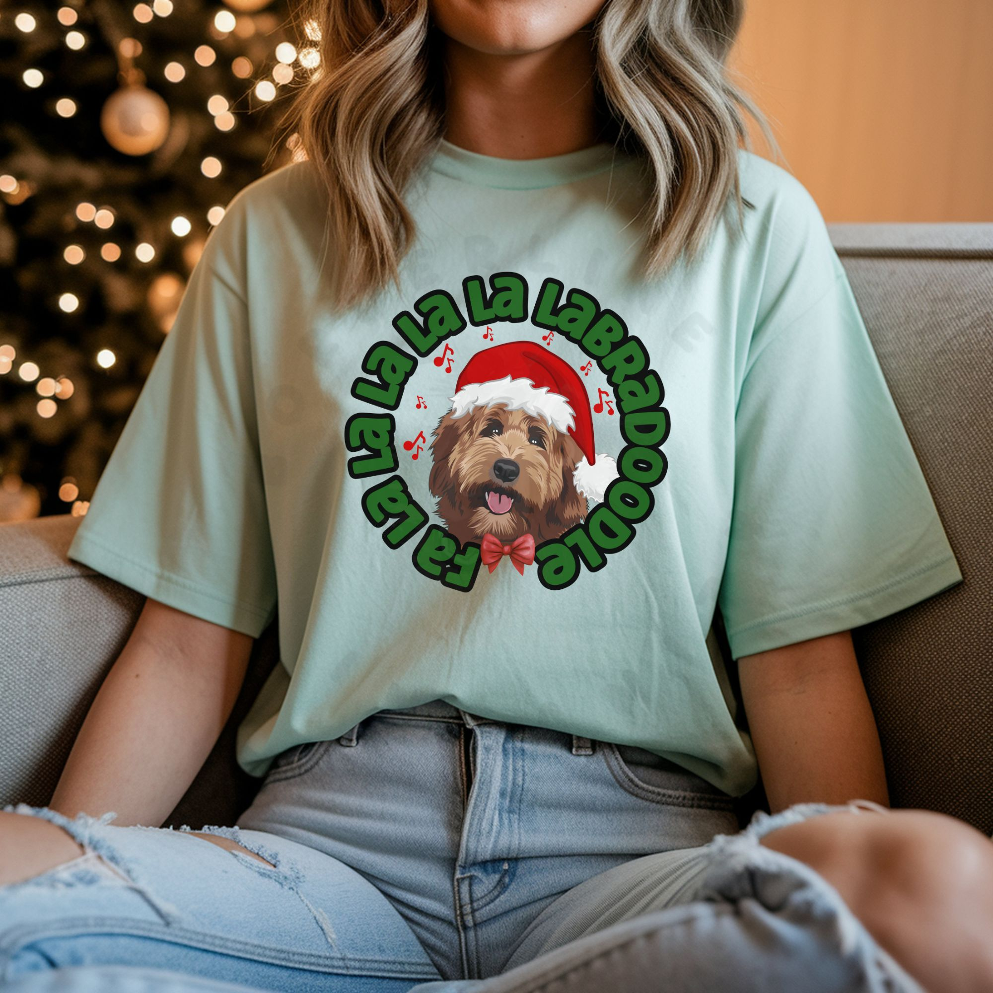 a woman sitting on a sofa in front of a christmas tree wearing a mint green tee featuring a graphic of a labradoodle wearing a santa hat and the words Fa la la la la la labradoodle