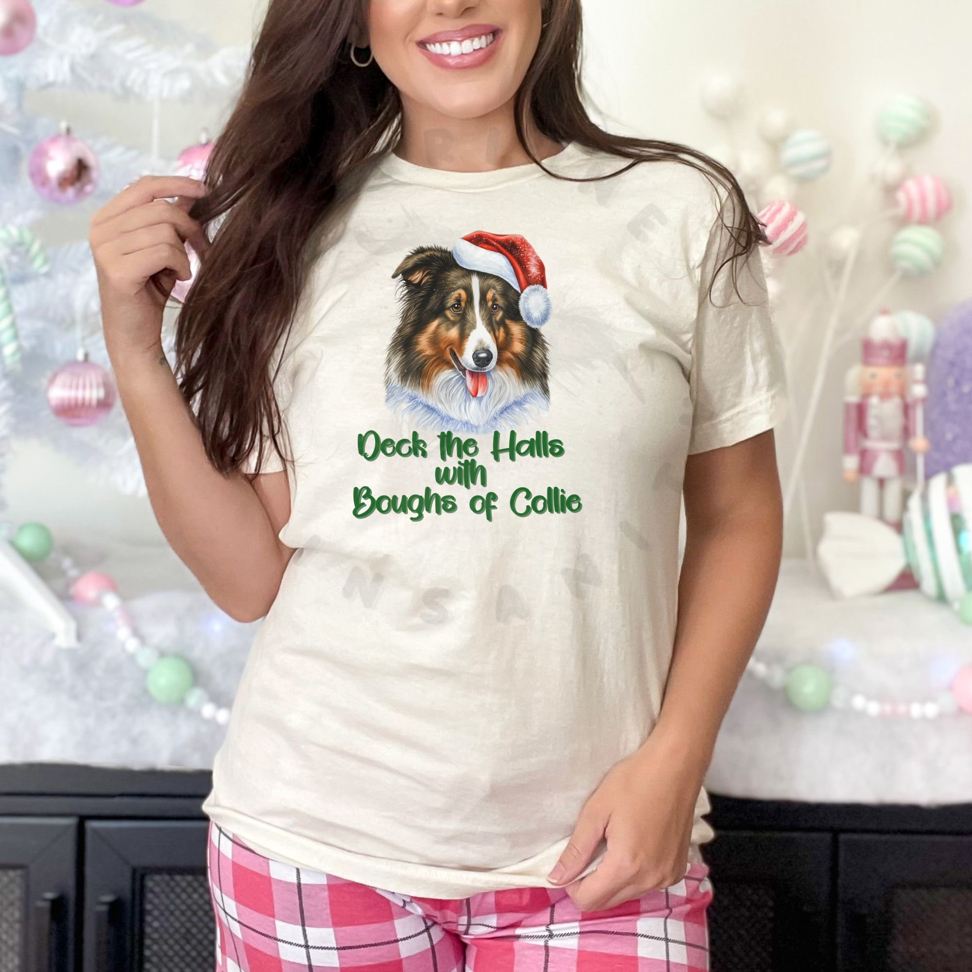 a woman wearing pink plaid pajama pants standing in front of a pastel christmas scene wearing an ivory (natural) t-shirt that features a rough collie wearing a santa hat and says Deck the halls with boughs of collie