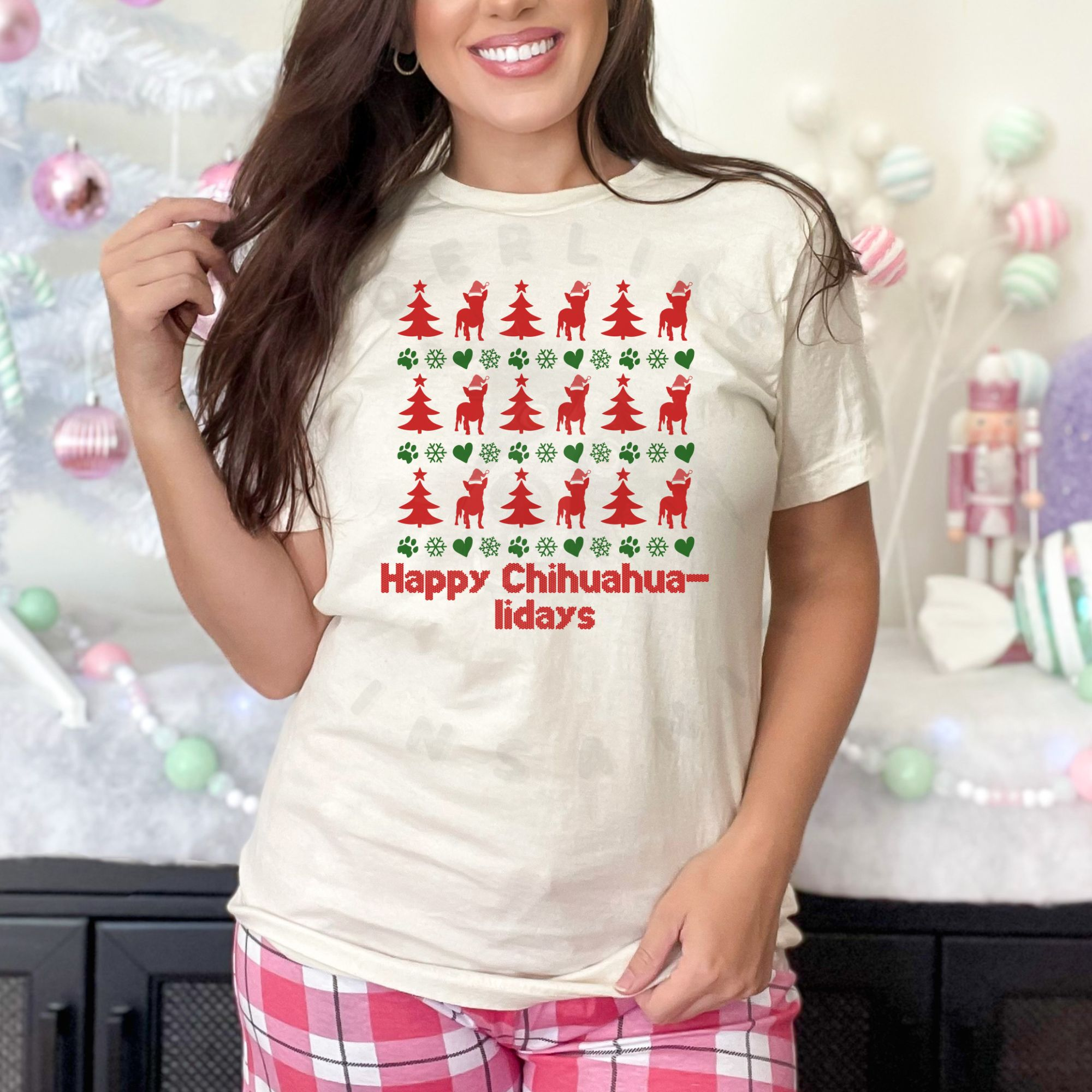 a woman wearing pink plaid pajama pants standing in front of a pastel christmas scene wearing an ivory (natural) t-shirt designed ugly christmas sweater style with rows of red christmas trees, green snowflakes, and chihuahuas wearing santa hats.