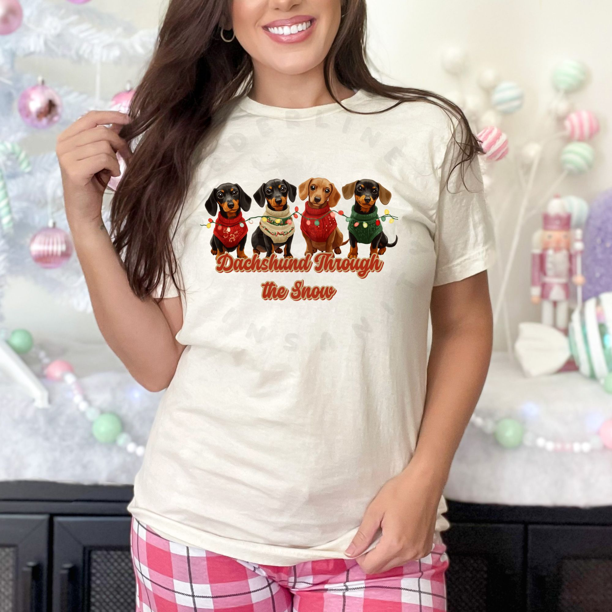 a woman in pink plaid pajama pants standing in front of a pastel christmas scene wearing an ivory (natural) dachshund through the snow t-shirt featuring 4 dachshunds wearing christmas sweaters and tangled in christmas lights