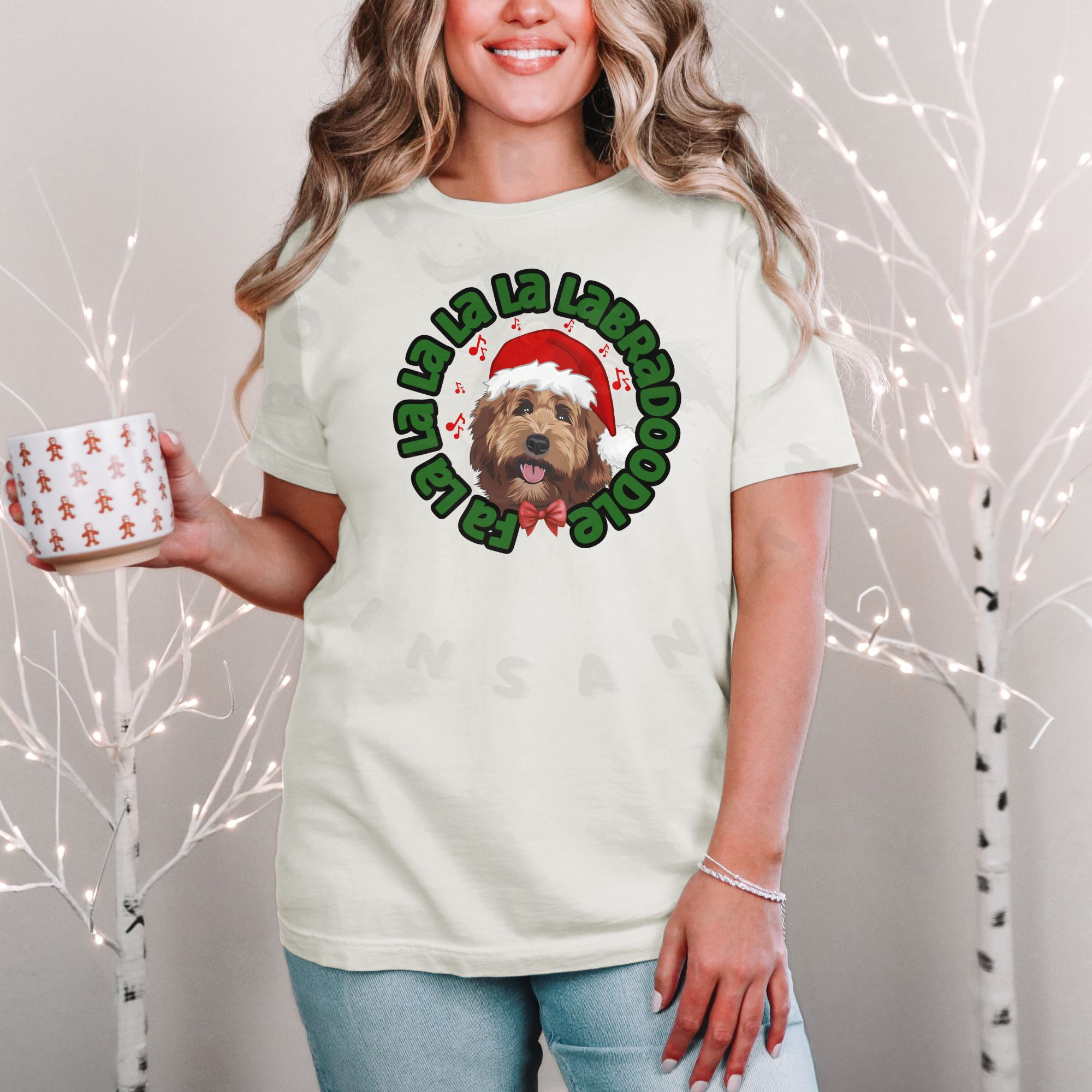 a woman standing in front of a christmas background holding a holiday mug wearing an ivory (natural) tee featuring a graphic of a labradoodle wearing a santa hat and the words Fa la la la la la labradoodle
