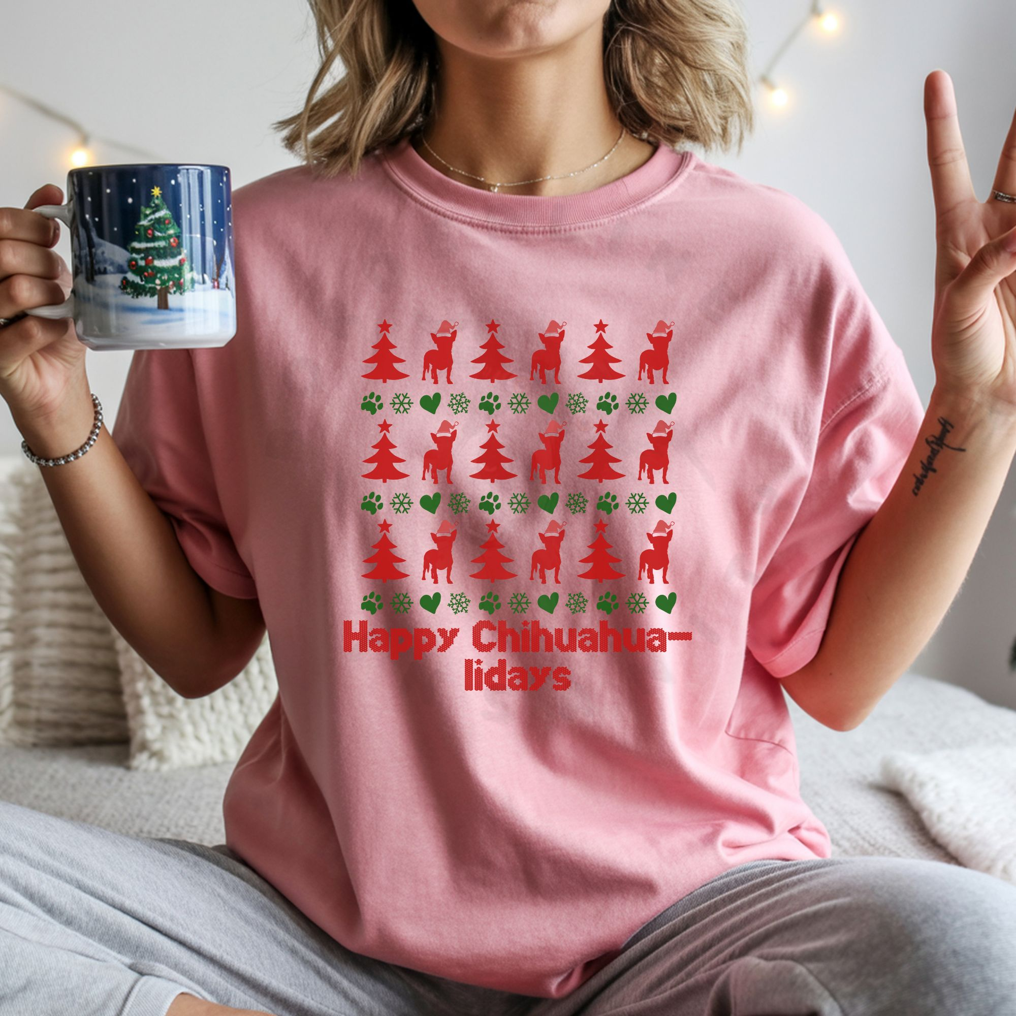 a woman holding a christmas mug wearing a pink t-shirt designed ugly christmas sweater style with rows of red christmas trees, green snowflakes, and chihuahuas wearing santa hats.