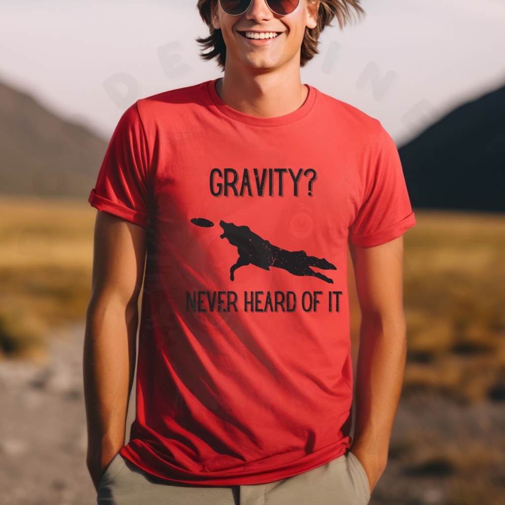 a man standing in the middle of a road in a desert wearing a red tee that says gravity? never heard of it with a silhouette of a dog leaping for a frisbee
