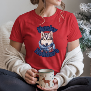 a woman sitting in front of a christmas tree holding a reindeer mug wearing a red tee that says husky holidays with a husky wrapped in a festive scarf