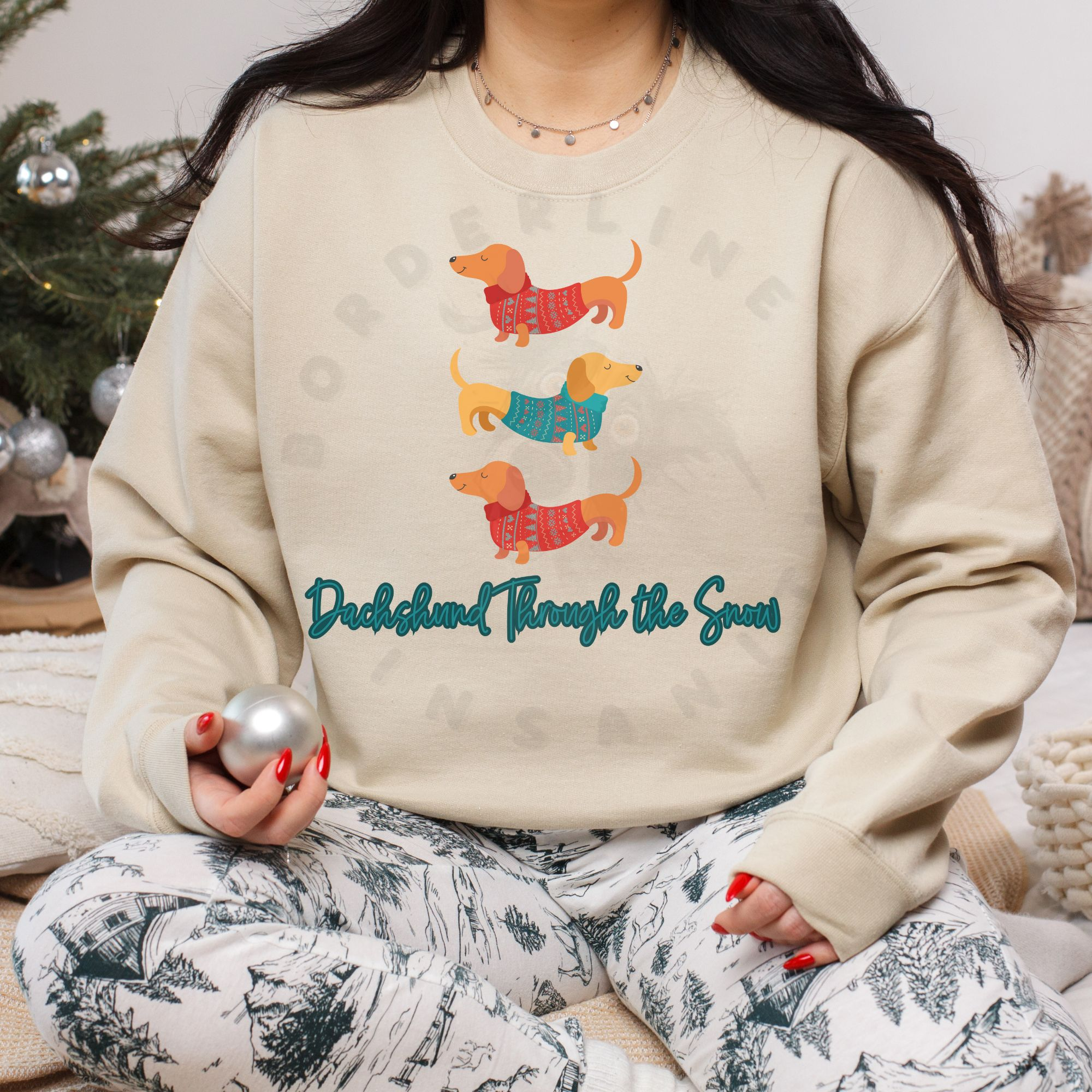 a woman sitting on the floor holding a silver ornament wearing a sand sweatshirt that says dachshund through the snow with a graphic of three dachshunds stacked wearing festive shirts.