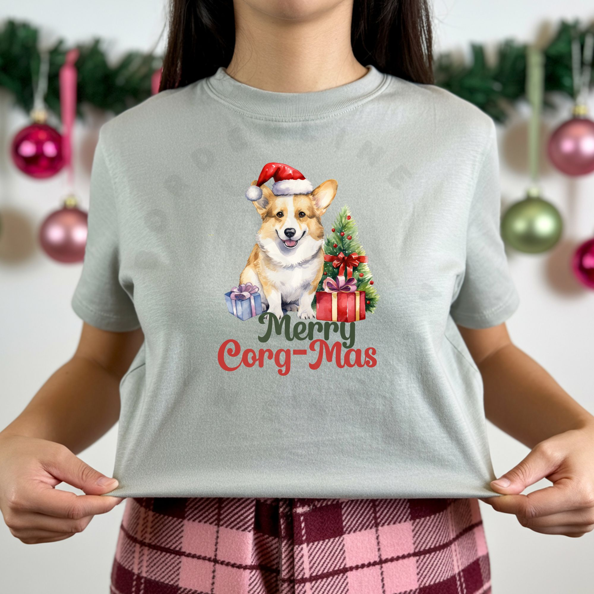a woman standing in front of an evergreen garland with christmas ornaments hanging from it wearing a silver t-shirt with a graphic of a corgi in a Santa hat with a Christmas tree and gifts that says Merry Corg-mas
