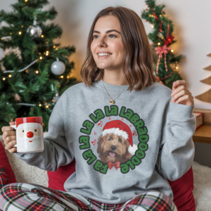 a woman sitting in front of a christmas tree holding a santa mug wearing a sport grey sweatshirt featuring a graphic of a labradoodle wearing a santa hat and the words Fa la la la la la labradoodle