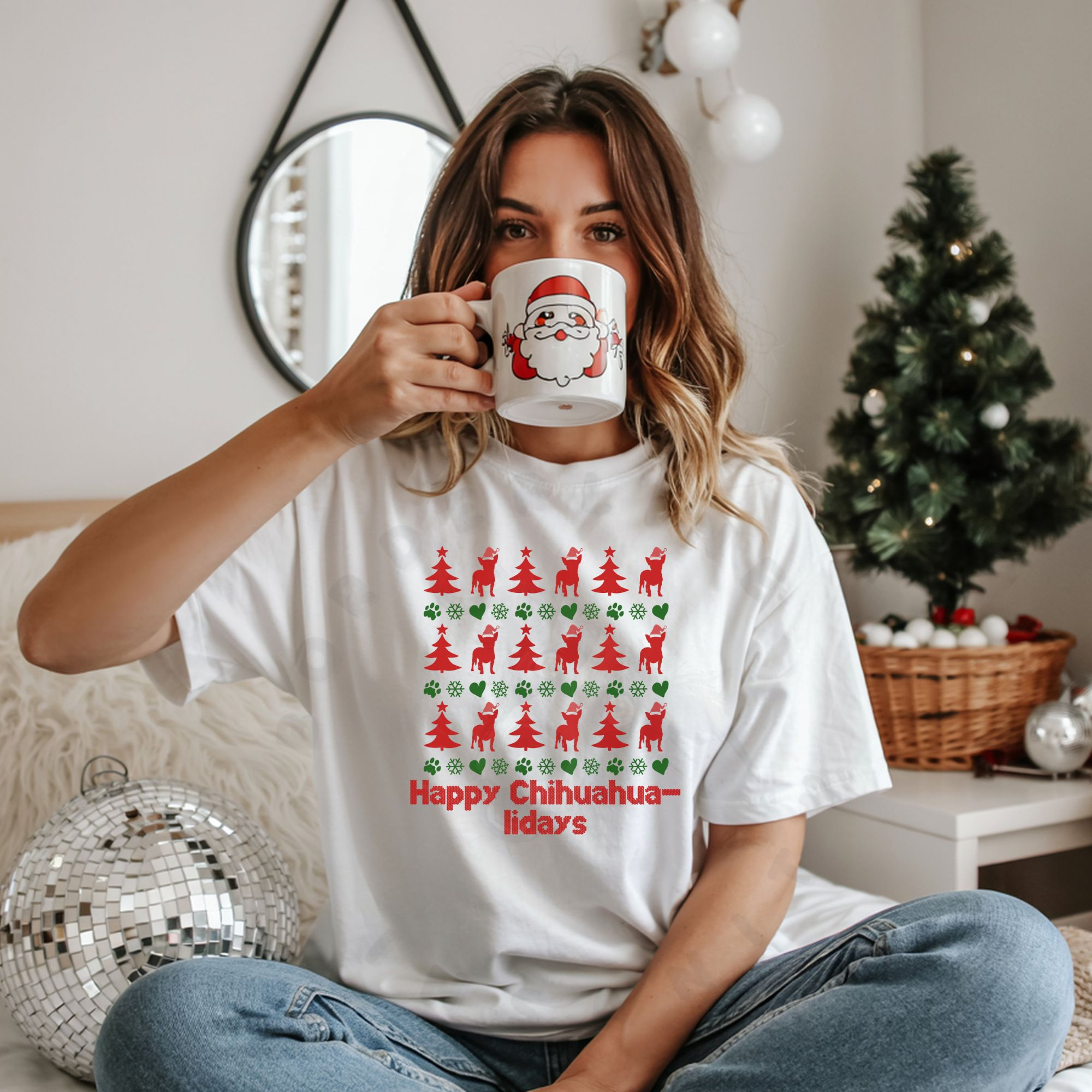 a woman sitting on a bed holding a christmas mug in front of her face wearing a white t-shirt designed ugly christmas sweater style with rows of red christmas trees, green snowflakes, and chihuahuas wearing santa hats.