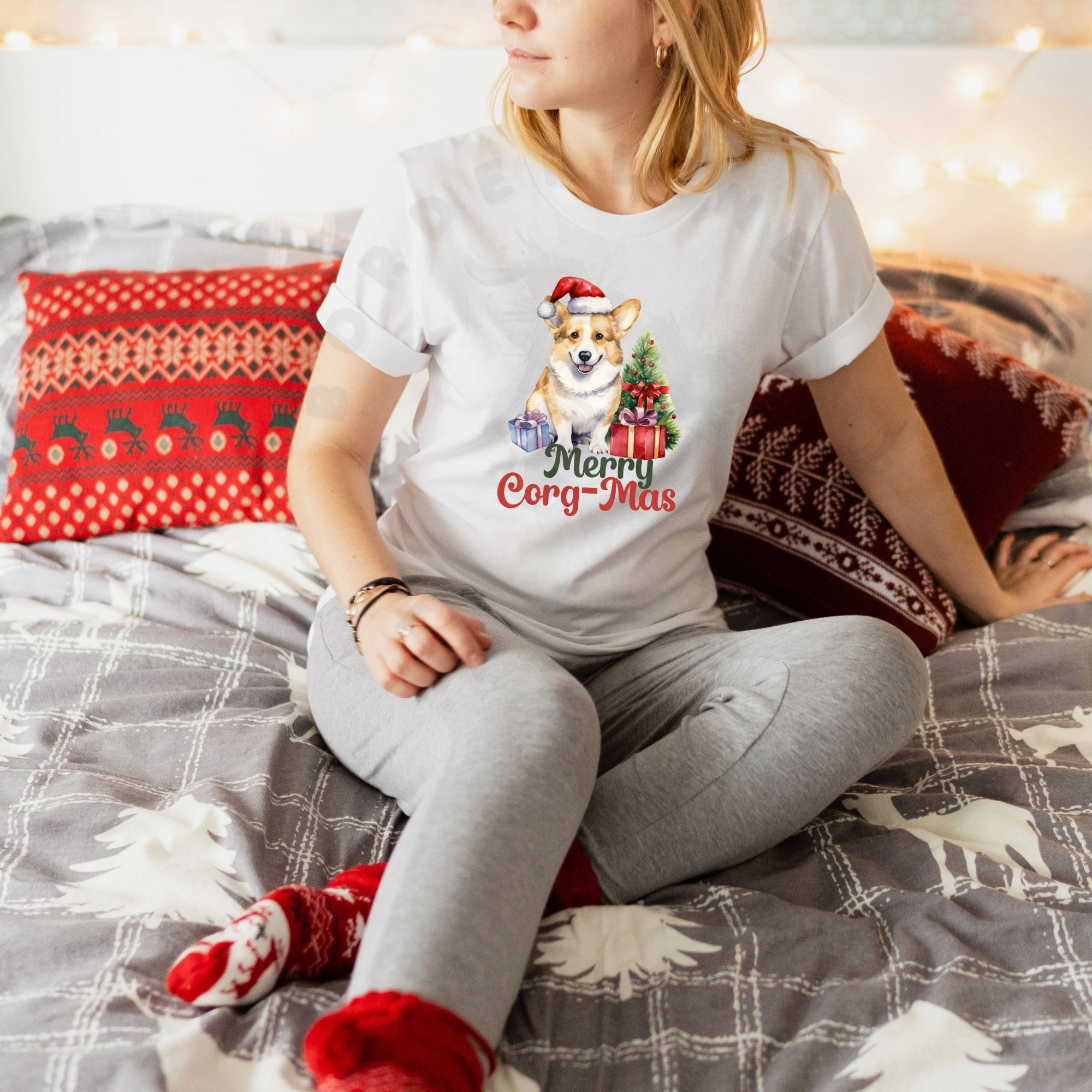 a woman sitting against christmas pillows wearing a white t-shirt with a graphic of a corgi in a Santa hat with a Christmas tree and gifts that says Merry Corg-mas