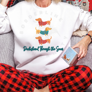 a woman in red buffalo plaid pajama pants sitting on a couch wearing a white sweatshirt that says dachshund through the snow with a graphic of three dachshunds stacked wearing festive shirts.