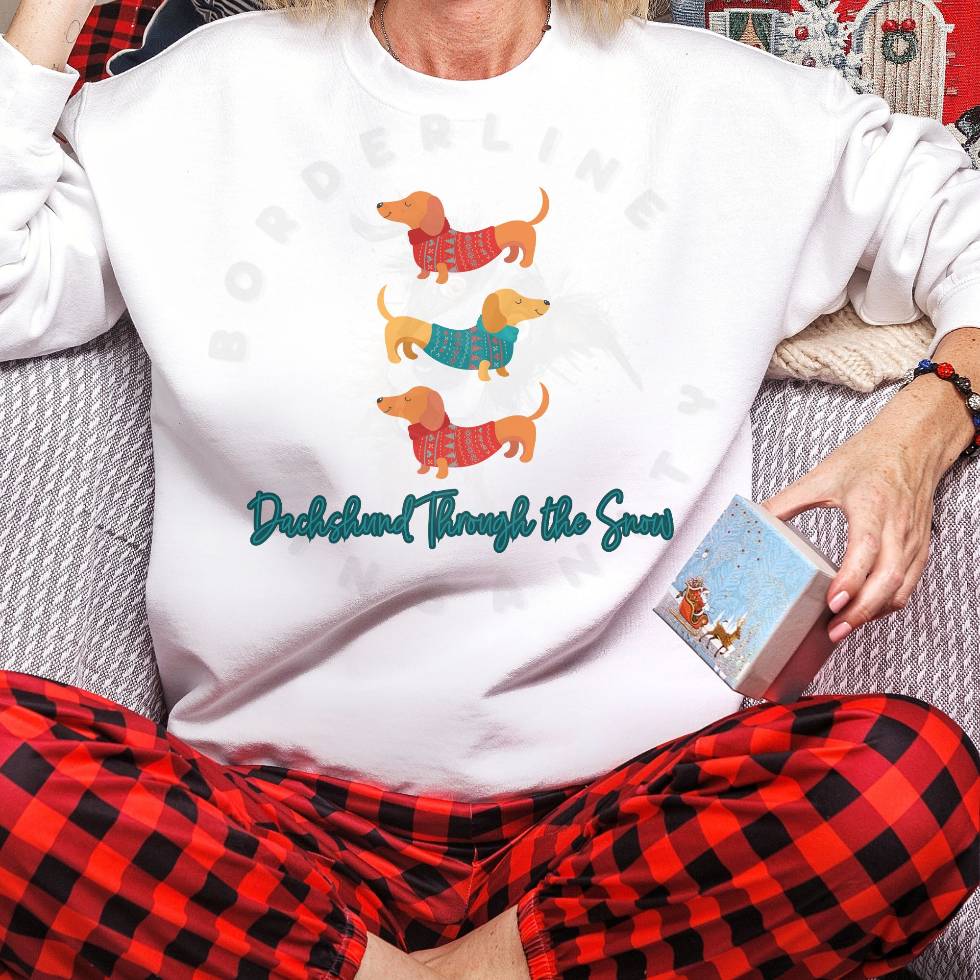a woman in red buffalo plaid pajama pants sitting on a couch wearing a white sweatshirt that says dachshund through the snow with a graphic of three dachshunds stacked wearing festive shirts.