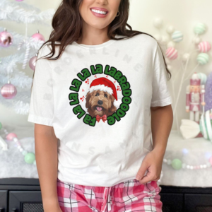 a woman wearing pink pajama pants standing in front of a pastel christmas background wearing a white tee featuring a graphic of a labradoodle wearing a santa hat and the words Fa la la la la la labradoodle