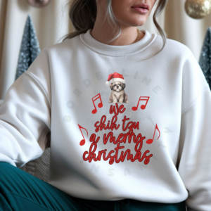 a woman sitting in front of christmas decorations wearing a white sweatshirt that says we shih tzu a merry christmas with a shih tzu wearing a santa hat at the top
