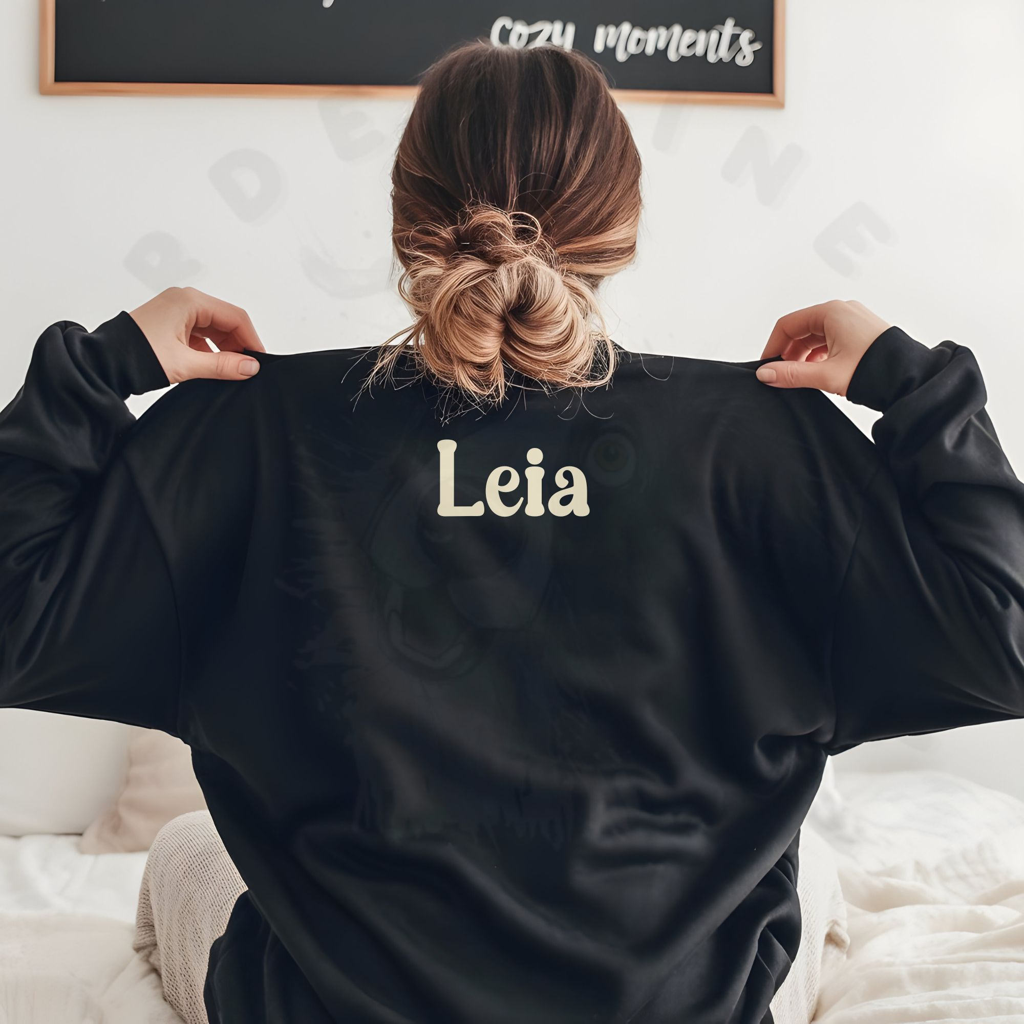 a woman sitting on a bed facing away from the camera wearing a black sweatshirt that says her name on the back