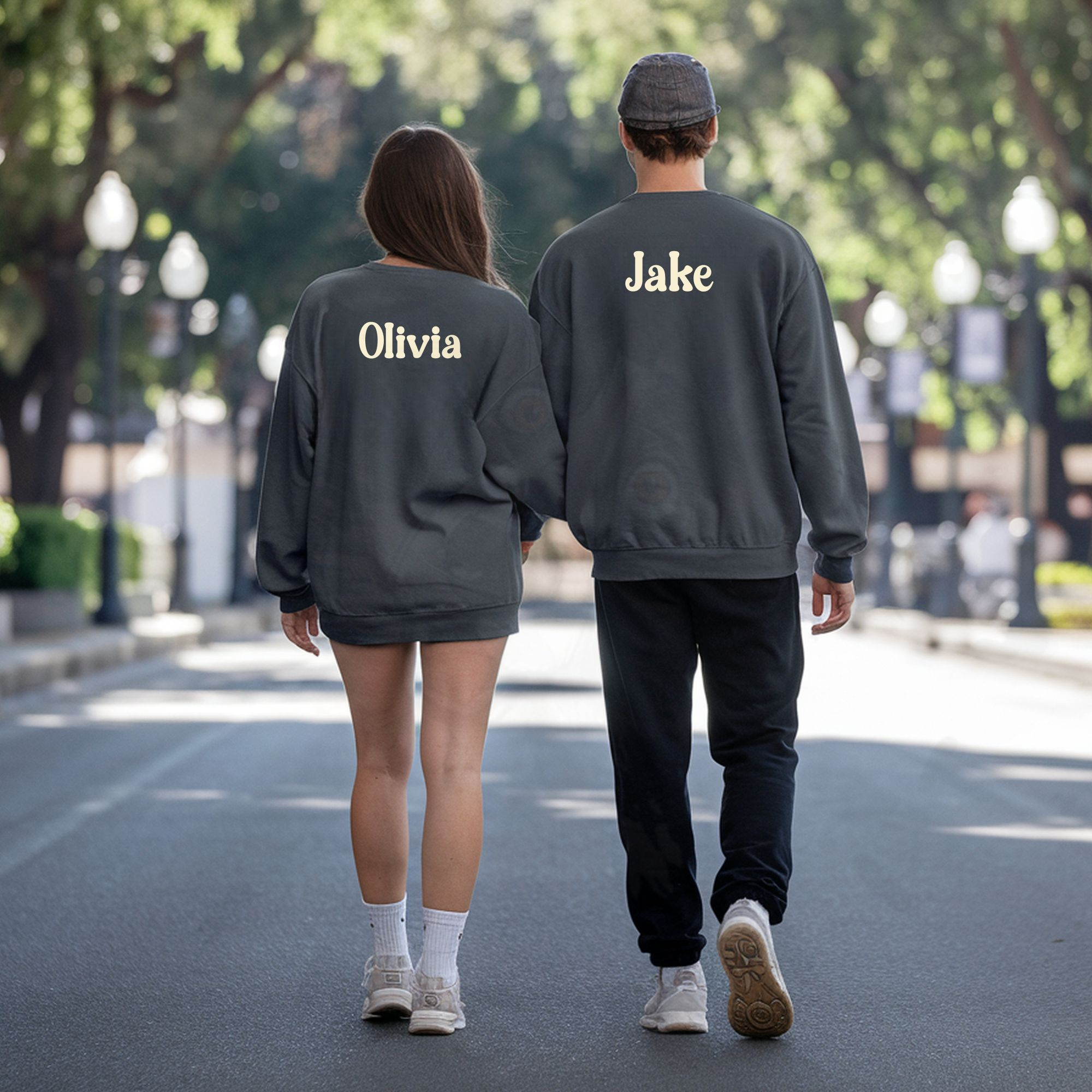 a man and woman walking in a park facing away from the camera wearing charcoal grey sweatshirts that have their names on the back