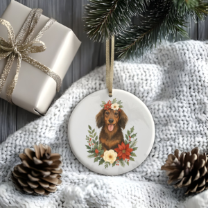 a stylized christmas scene of a package, pine cones, greenery, and a blanket with a round ceramic christmas ornament featuring a graphic of christmas flowers and greenery surrounding a dachshund