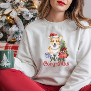 a woman holding a green christmas mug and sitting in front of a christmas tree wearing a white sweatshirt with a graphic of a corgi in a santa hat with a christmas tree and gifts that says Merry Corg-mas