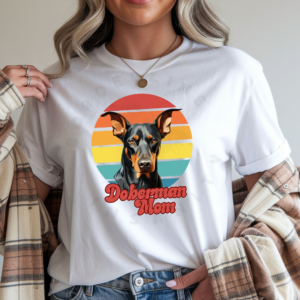a woman wearing a white tee featuring a retro sunset design with a doberman and the words doberman mom in retro script