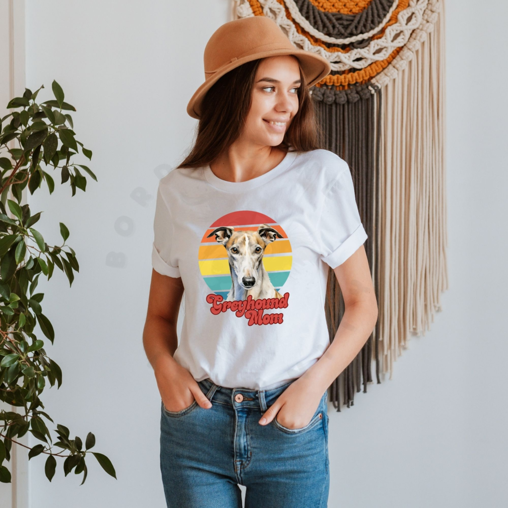 a woman wearing a white tee featuring a retro sunset design with a greyhound and the words greyhound mom in retro script