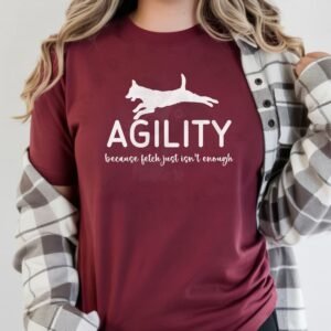 a woman wearing a plaid flannel overshirt wearing a maroon tee with the words Agility because fetch just isn't enough and a silhouette of a jumping dog
