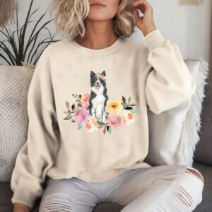 a woman on a sofa wearing a sand colored crewneck sweatshirt featuring an image of a border collie in watercolor flowers