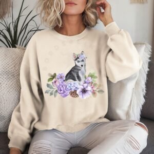 a woman on a sofa wearing a sand colored crewneck sweatshirt featuring an image of a husky puppy in watercolor flowers